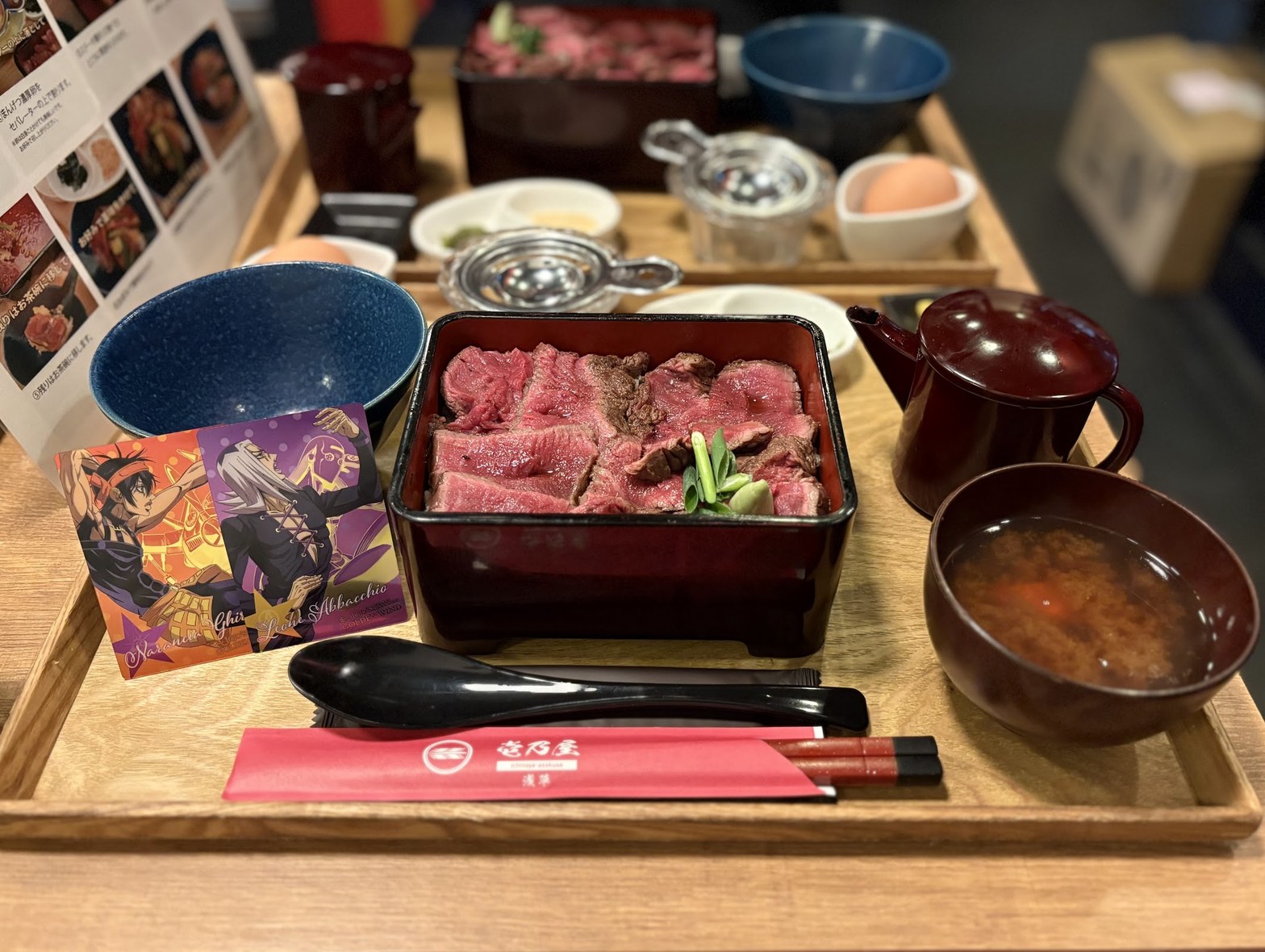 漆塗りお重に盛られたレアステーキと壱乃屋ロゴ入り赤い箸袋、アニメコラボカードが並ぶテーブル